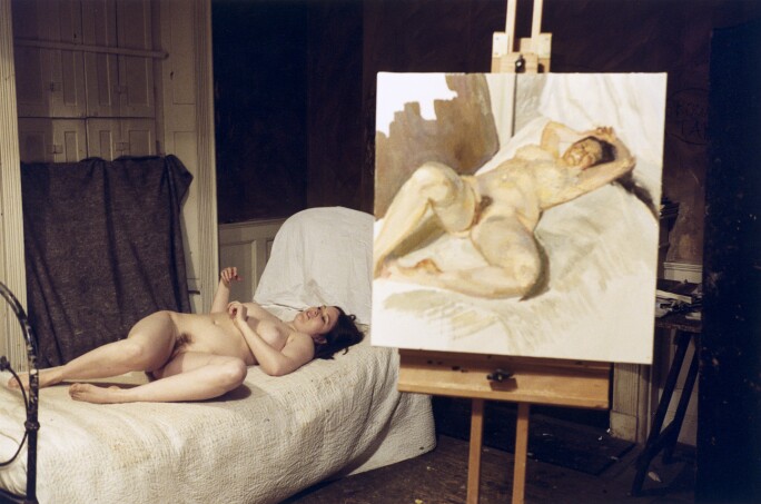 A nude white woman with dark hair reclines on a bed with dirty white sheets. A painting of her is on an easel to the right of the bed.
