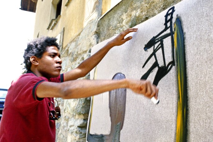 Photograph of Basquiat painting an outdoor wall in St. Moritz, Switzerland. He is wearing a
