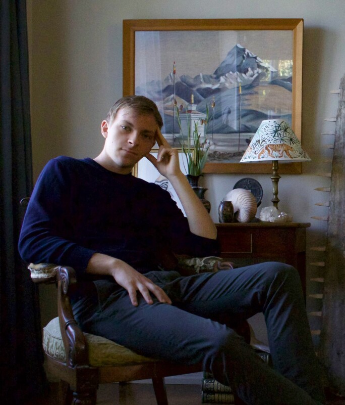 A blond young man seated in a chair with his head resting on his hand.