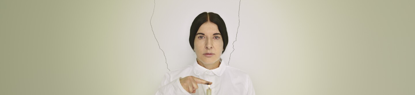 Marina Abramovic dressed in all white holds her finger, covered in soot, over a lit green candle. 