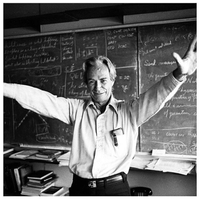 Richard Feynman in the classroom with his arms open and extended and standing in front of a blackboard covered in formulas.