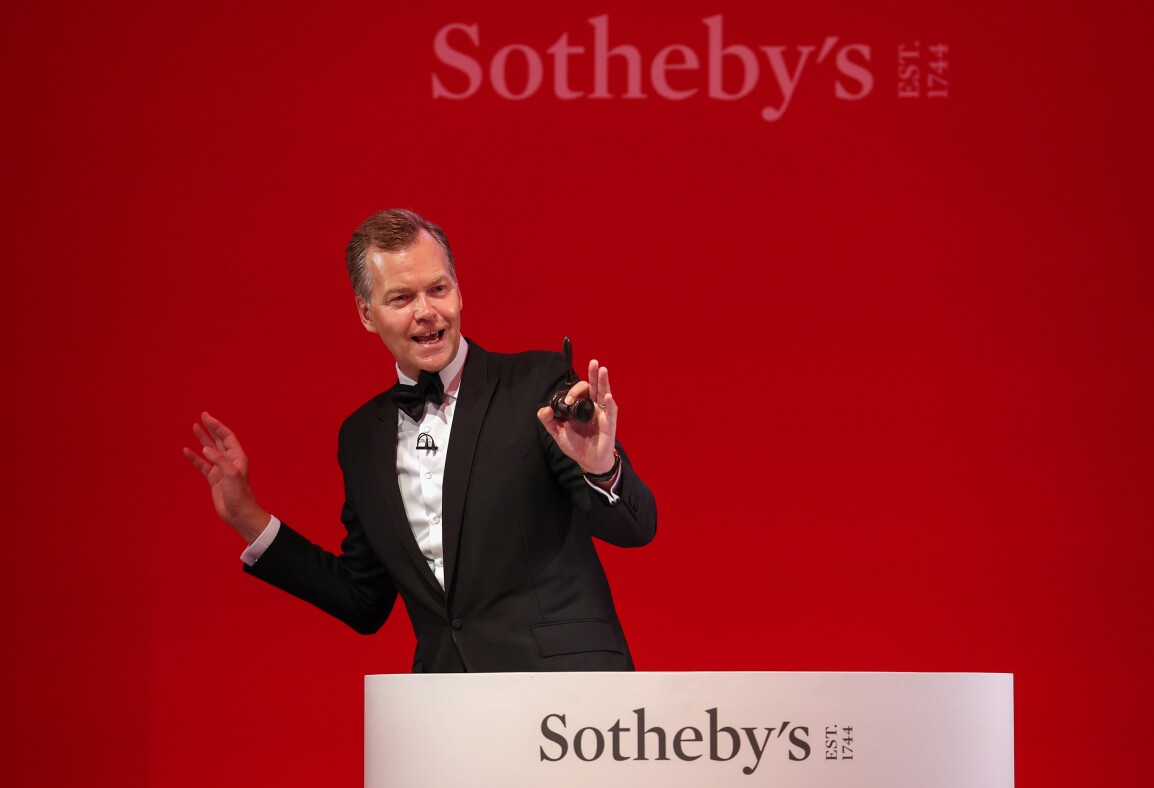 Sotheby's auctioneer Oliver Barker stands behind a rostrum holding a gavel. He's wearing a tuxedo and standing in front of a bright red wall.