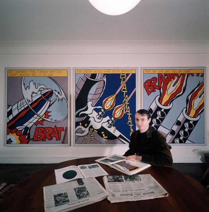The American painter and exponent of Pop Art, Roy Lichtenstein, is sitting at a table and is attentively looking through some magazines; over his shoulder some of his artwork. 1964.