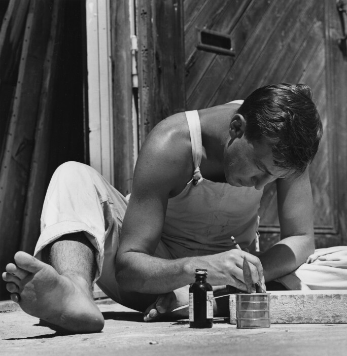 Black-and-white photograph of man drawing on board, with paintbrush, paint pot and ink beside him