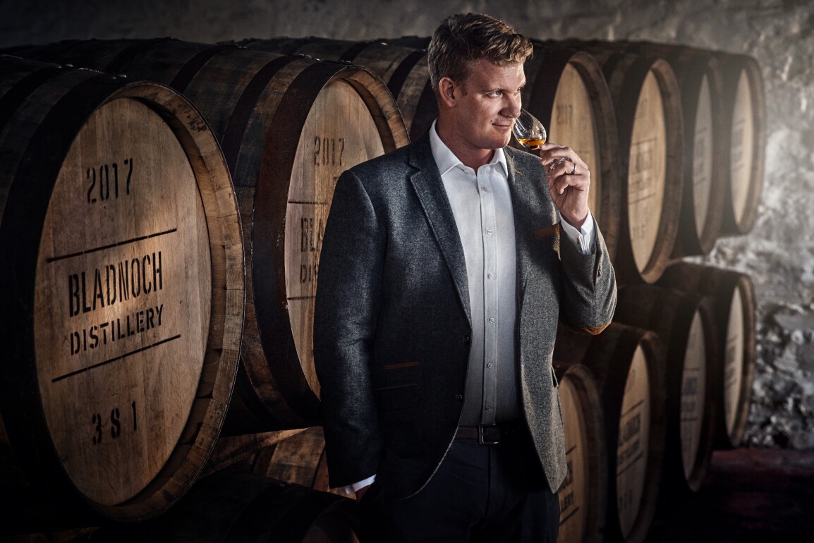 Man smelling whisky in front of barrels with "Bladnock Distillery" printed on the front