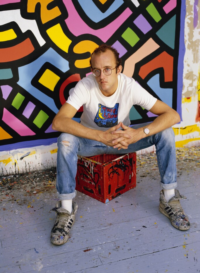 Keith Haring seated on a milk crate in front of a canvas.